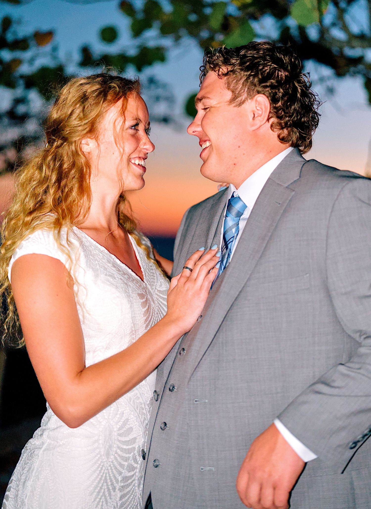 Happy bride and groom gazing at each other at sunset, she wears white gown, he wears gray suit.