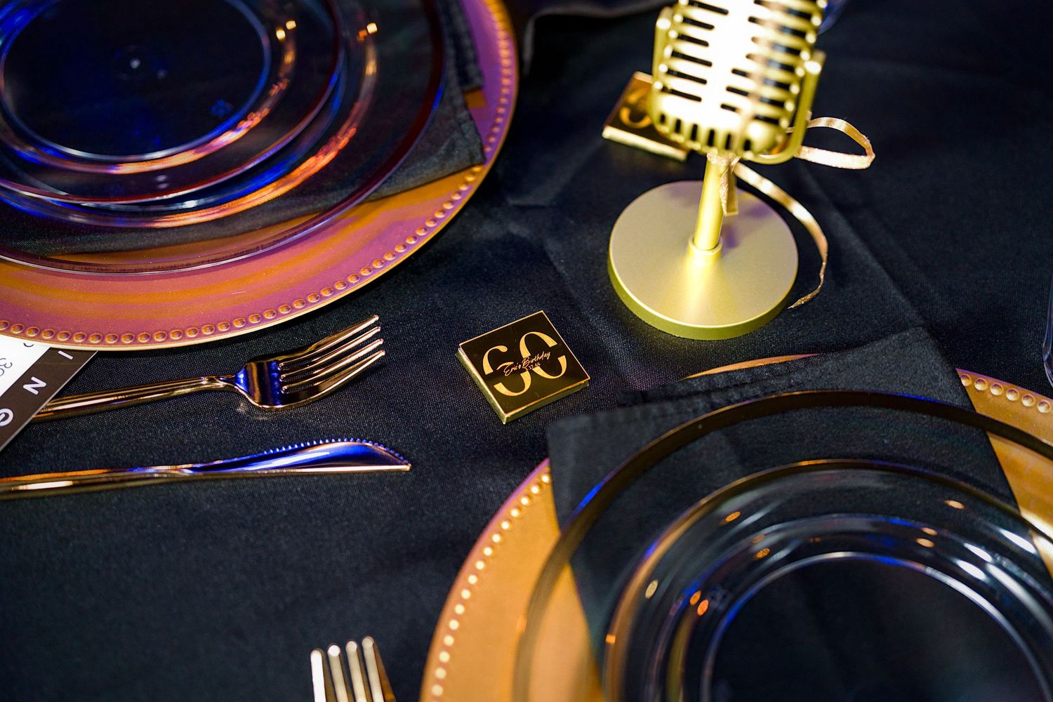Elegant event table setting with blue plates, gold cutlery, and a decorative gold microphone centerpiece on black linen.