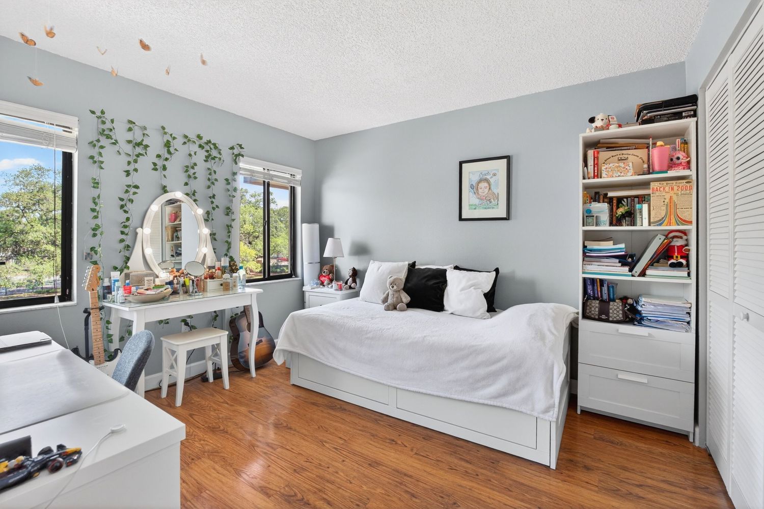 Bright bedroom with hardwood floors, white bed, vanity desk, and bookshelf, decorated with trailing ivy wall plants.