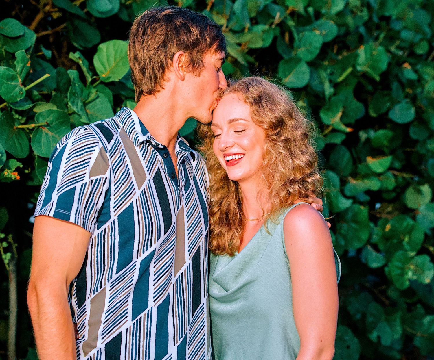 A young couple sharing a romantic kiss outdoors, surrounded by lush green tropical foliage.