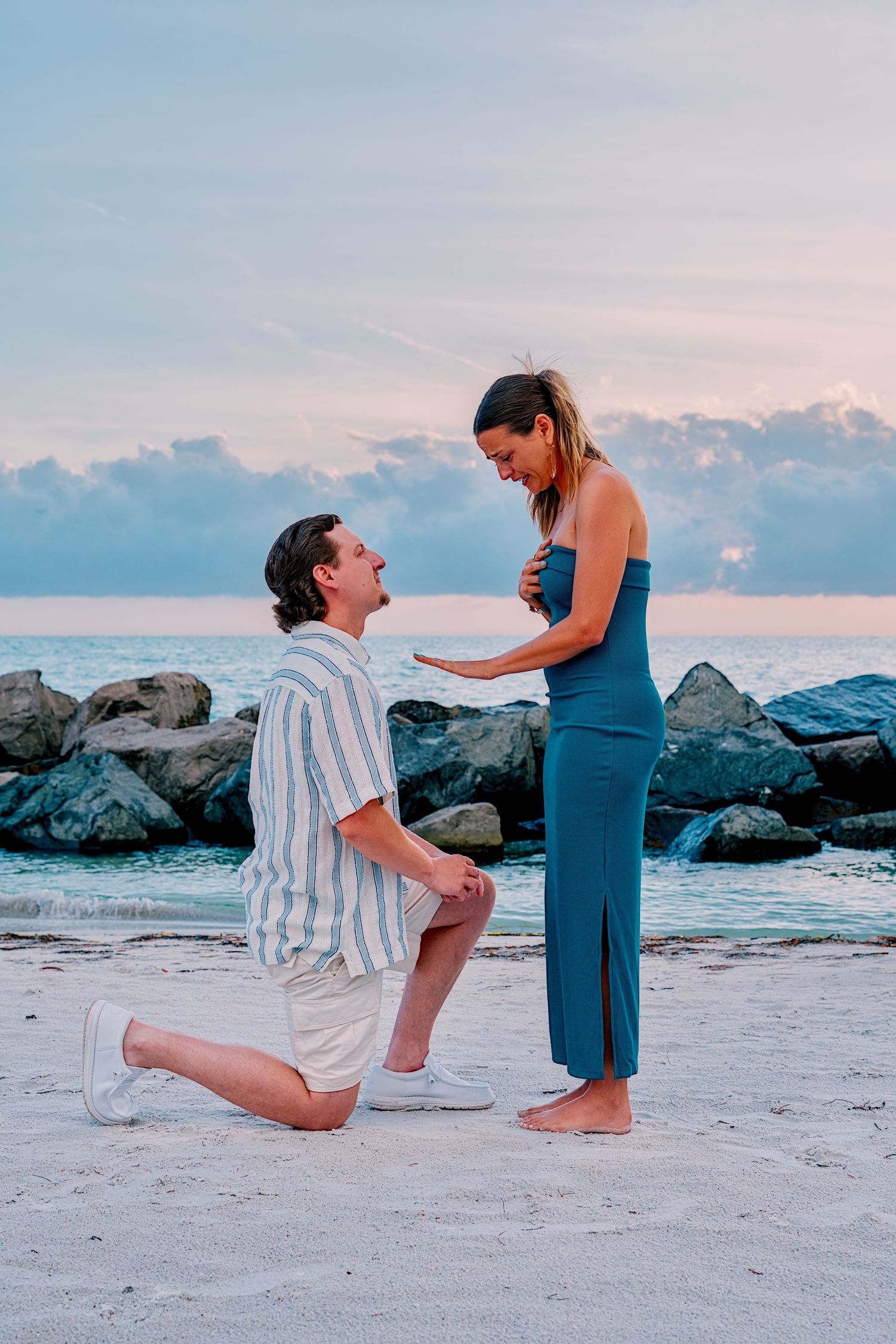 Man proposes on one knee to woman in blue dress on sandy beach at sunset with ocean backdrop.