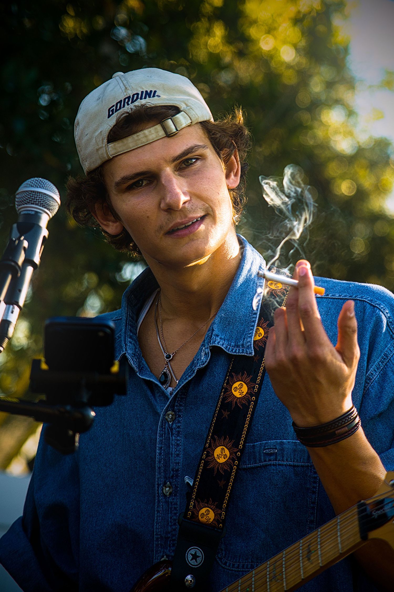 Young male musician with backward cap holds cigarette while playing guitar near microphone outdoors.