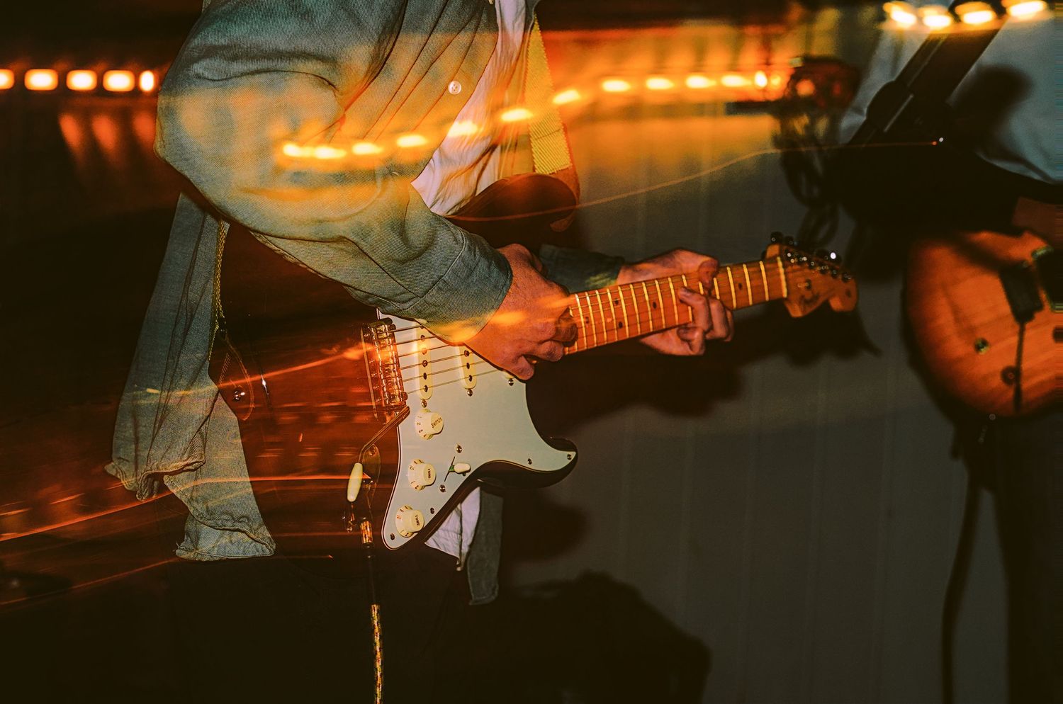 Musician playing a white Fender Stratocaster electric guitar on stage with warm bokeh lights in background.