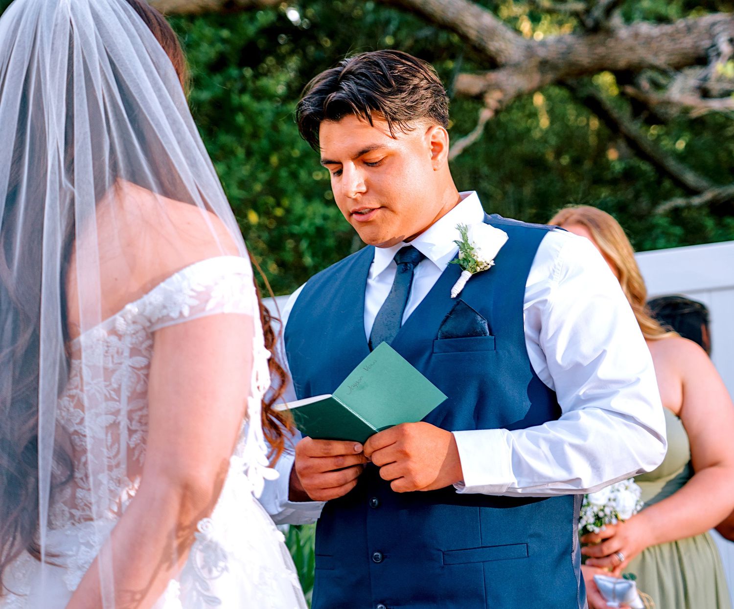 Groom in blue vest reading vows from a green card during an outdoor wedding ceremony.