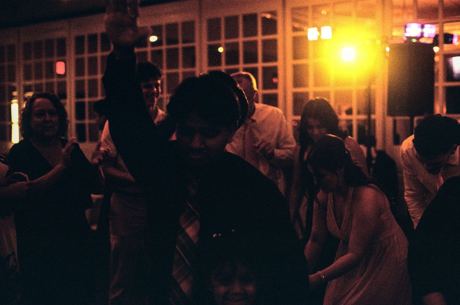 Guests dancing at a dimly lit indoor party with warm orange lighting and large windows in the background.
