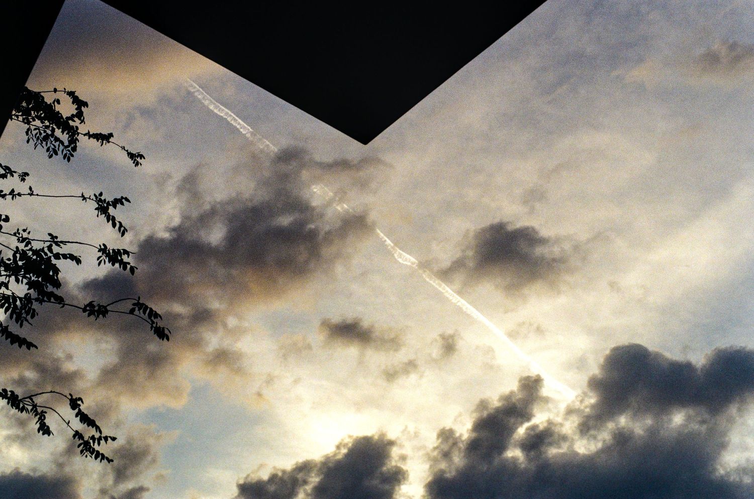 Airplane contrail streaking across dramatic cloudy sky at dusk, viewed through geometric opening with tree silhouette.