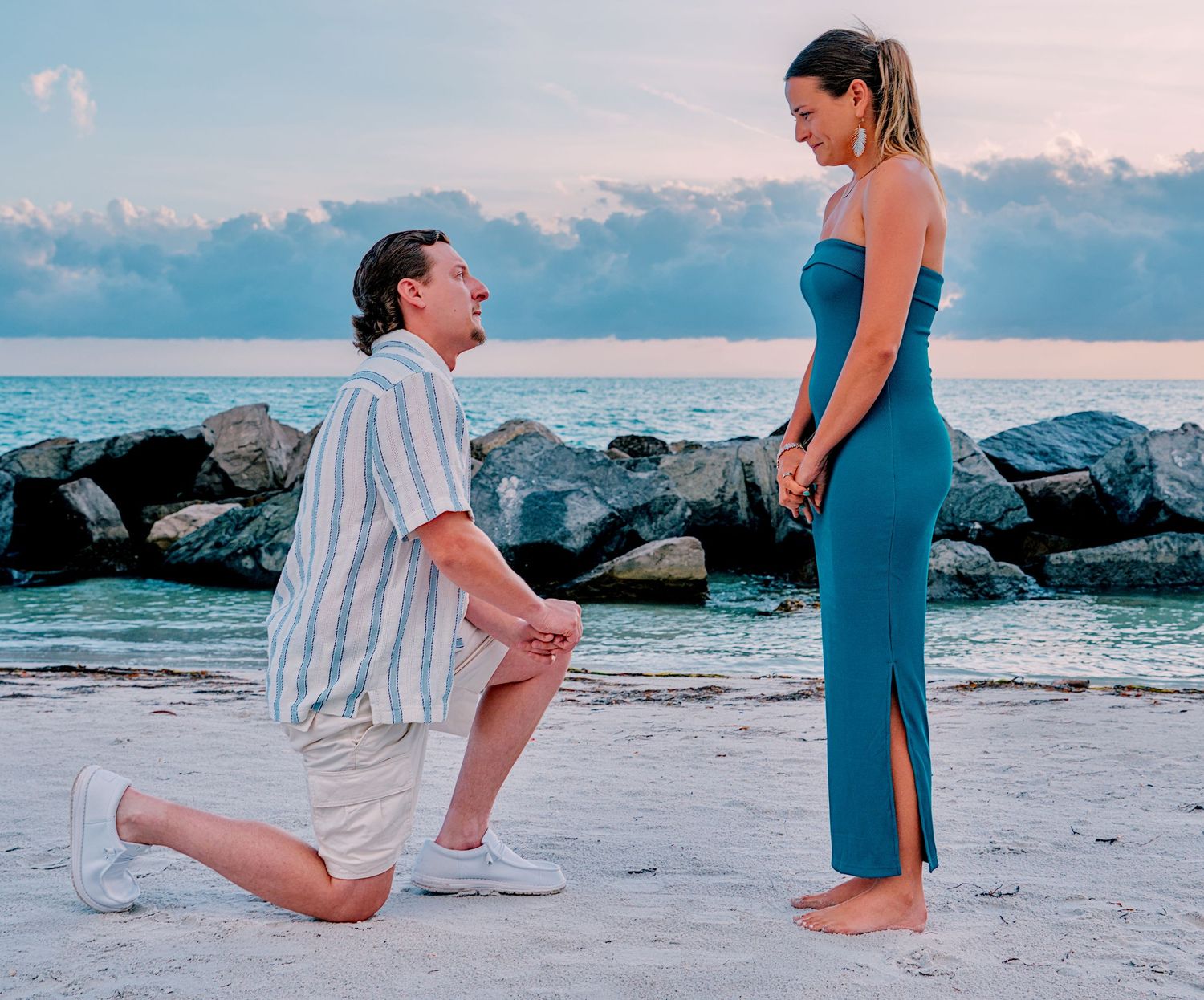 Man proposing on one knee to woman in teal dress on a sandy beach at sunset with rocky shoreline.
