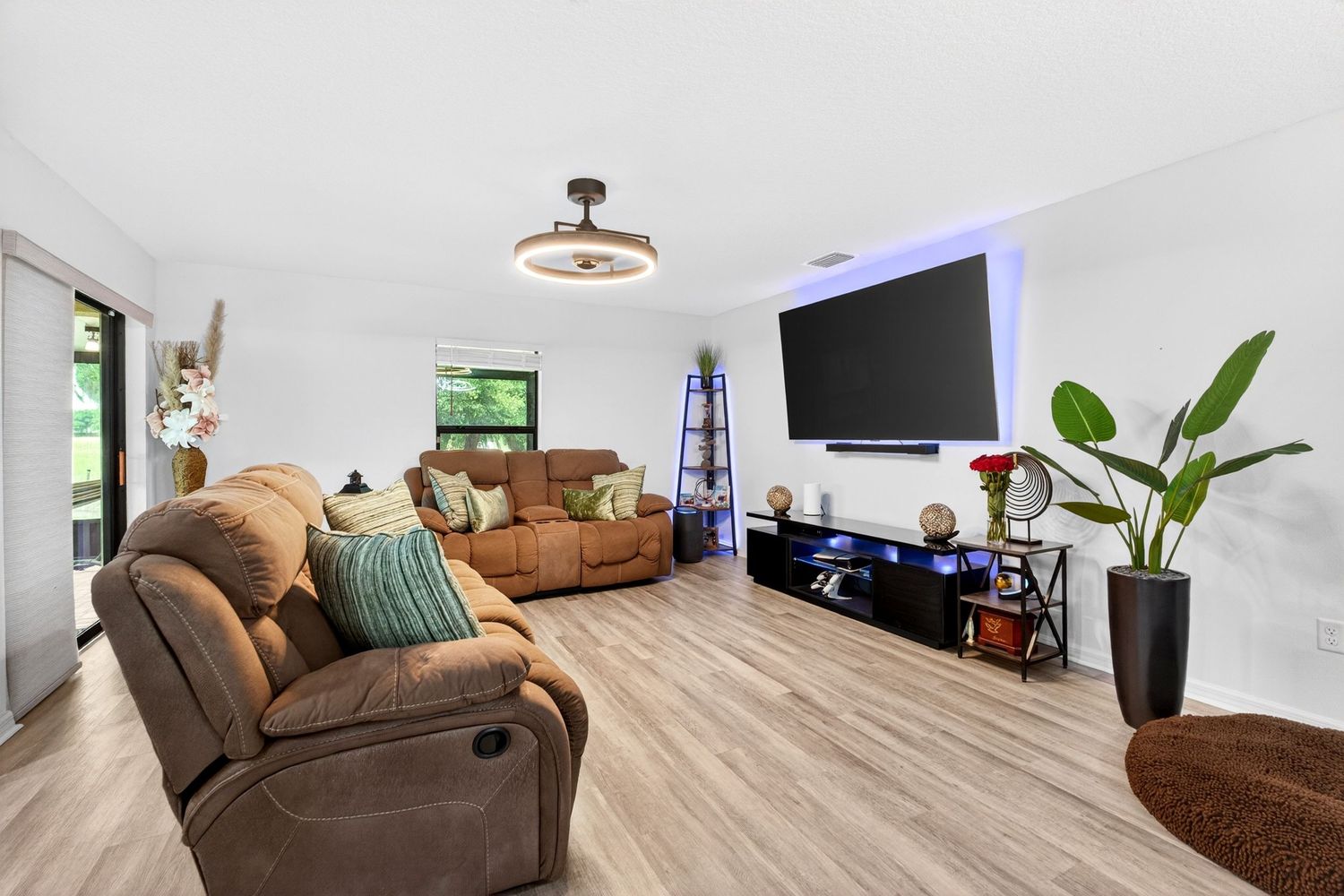 Bright living room with brown recliner sofas, light wood floors, large wall-mounted TV, and modern ceiling fan.