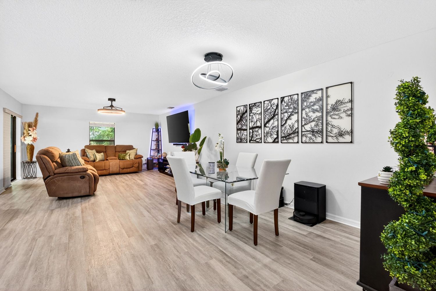 Open-concept living and dining room with light hardwood floors, white walls, modern furniture, and decorative wall art.