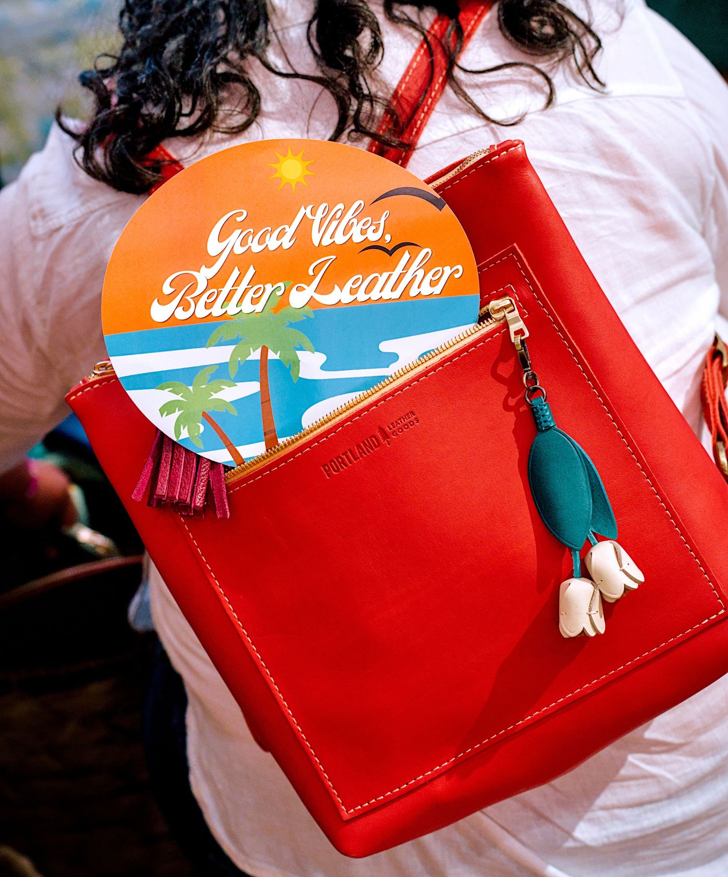 Red leather backpack with tassel charm and 'Good Vibes, Better Leather' tropical sign, worn by a person outdoors.