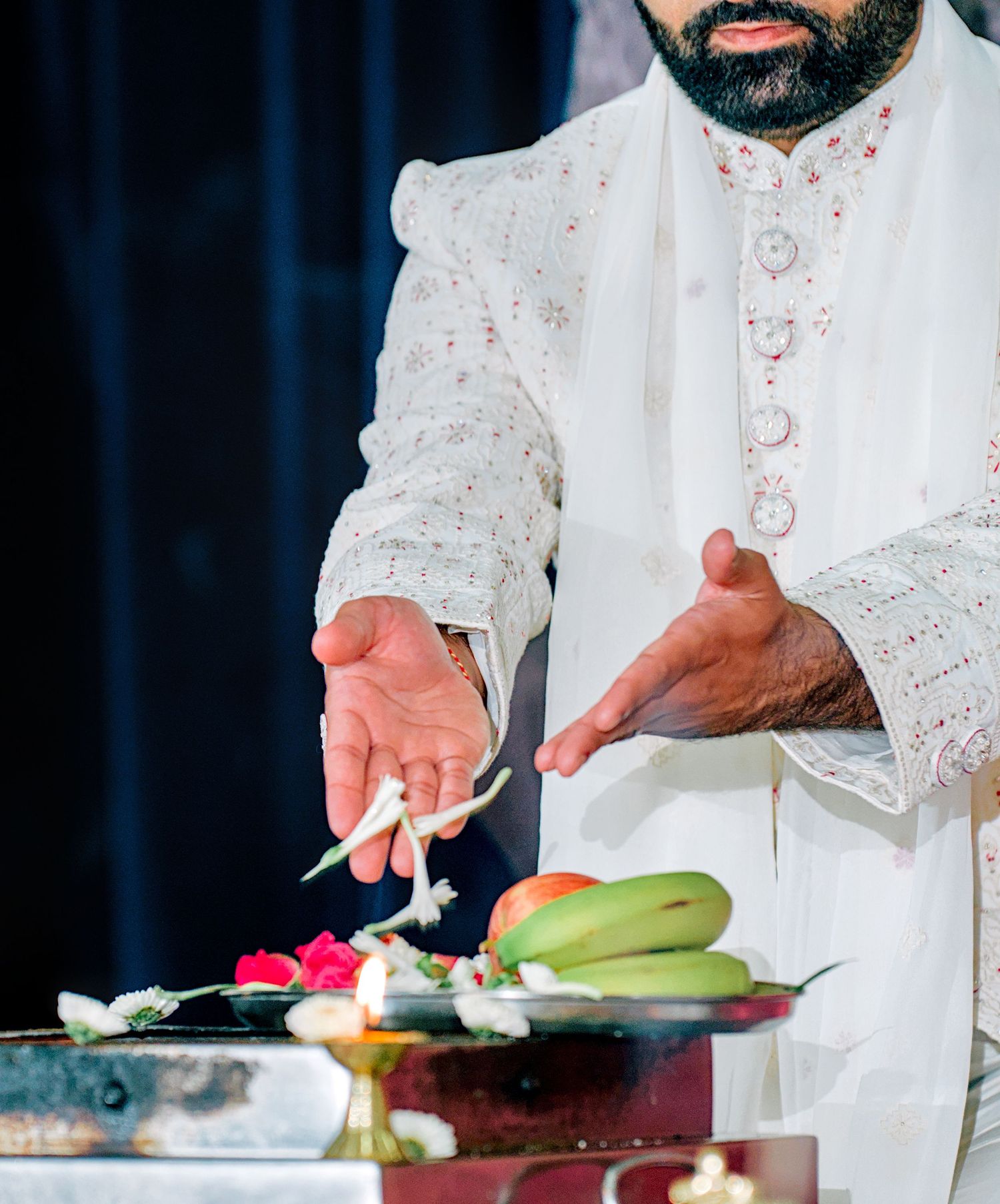 Man in white embroidered sherwani performing Hindu wedding ritual with flowers and offerings on decorated tray.