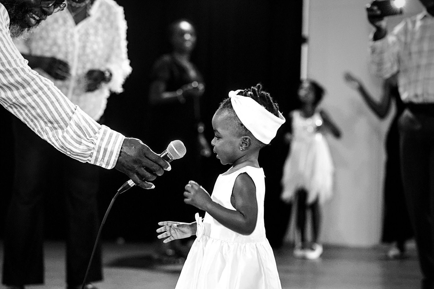 Black and white photo of a toddler girl in white dress being offered a microphone.
