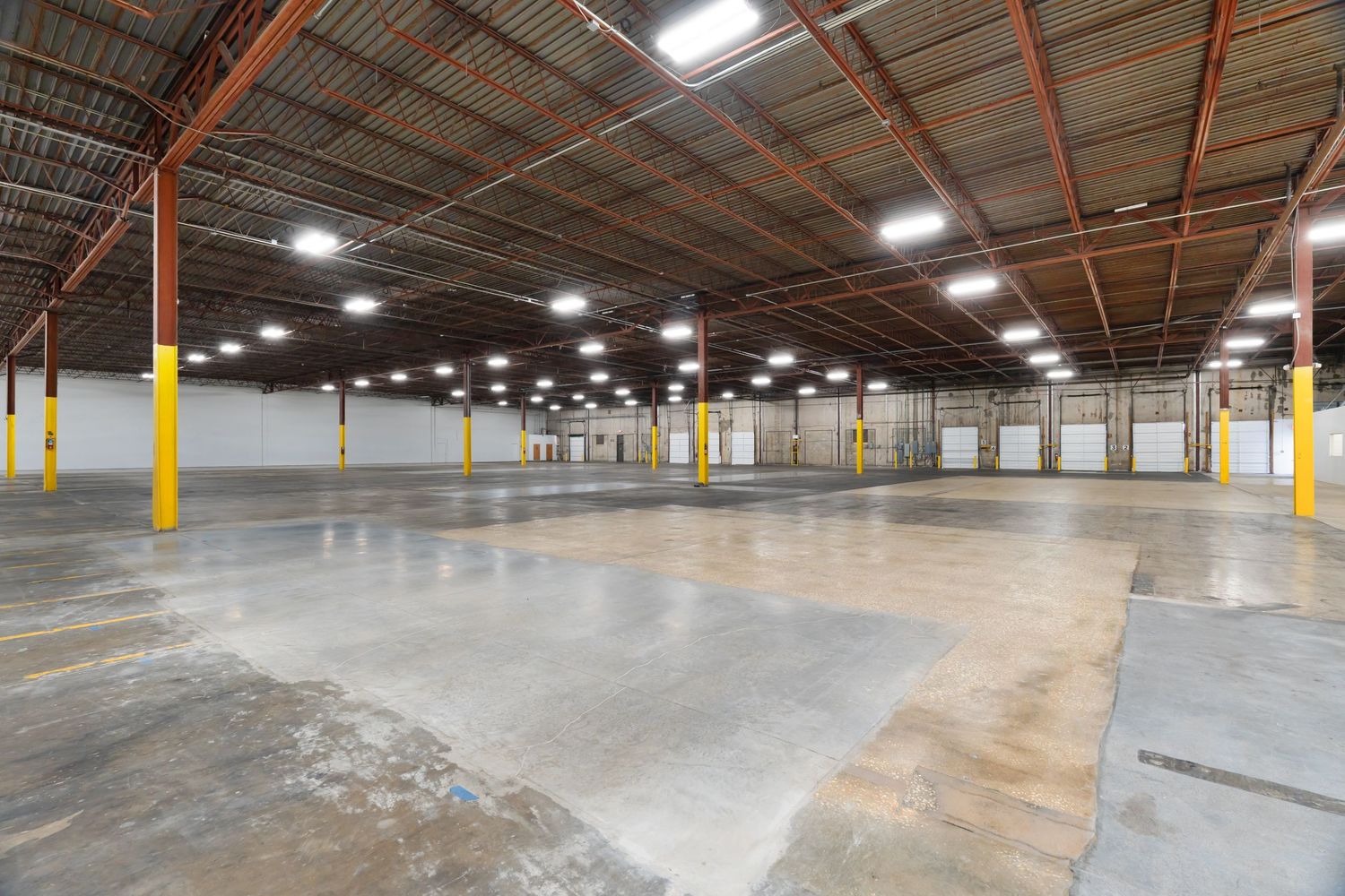 Large empty industrial warehouse interior with concrete floor, metal support beams, and bright overhead lighting.