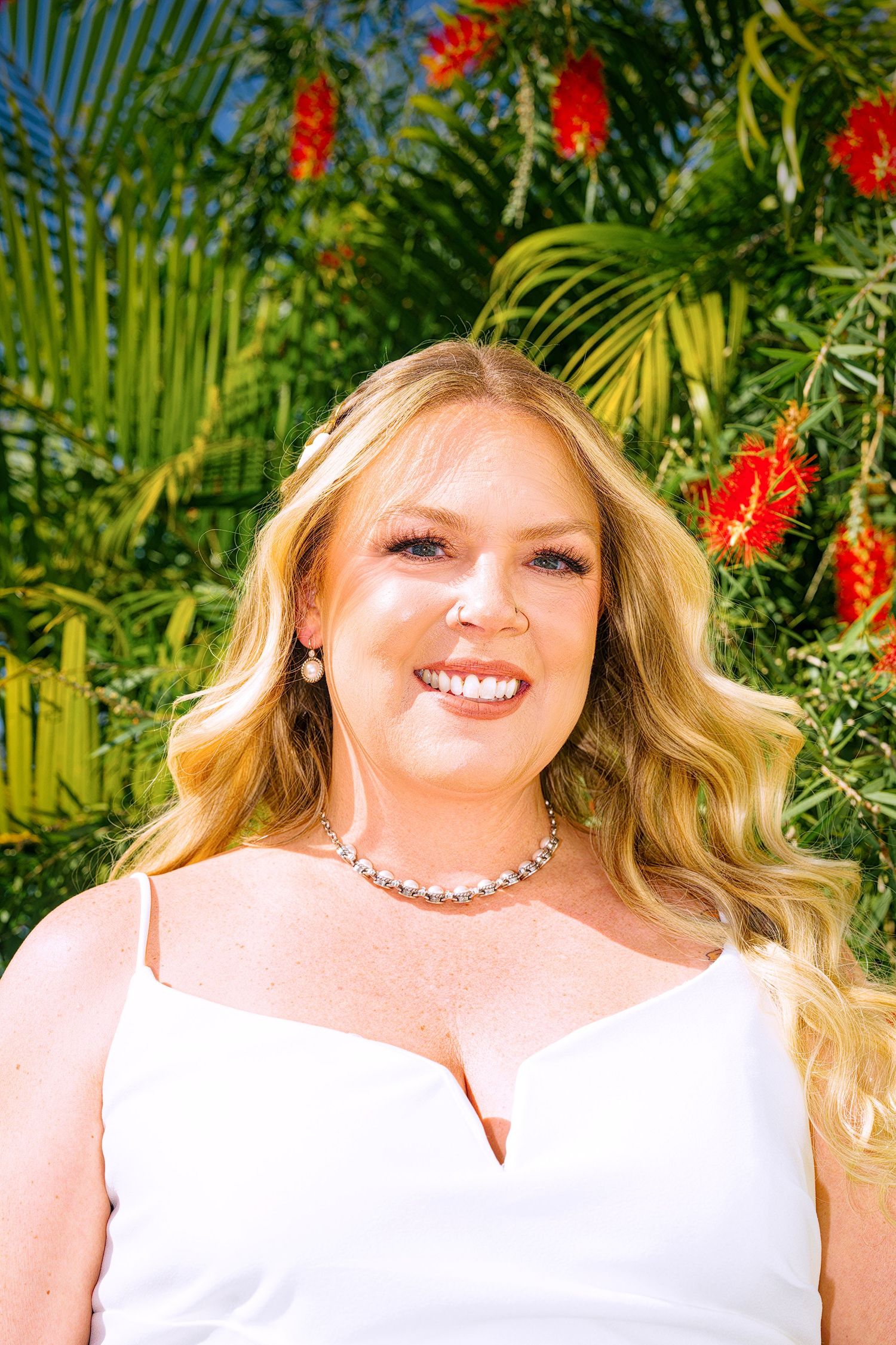 A smiling woman in a white dress poses against a tropical background with red flowers and palm fronds.