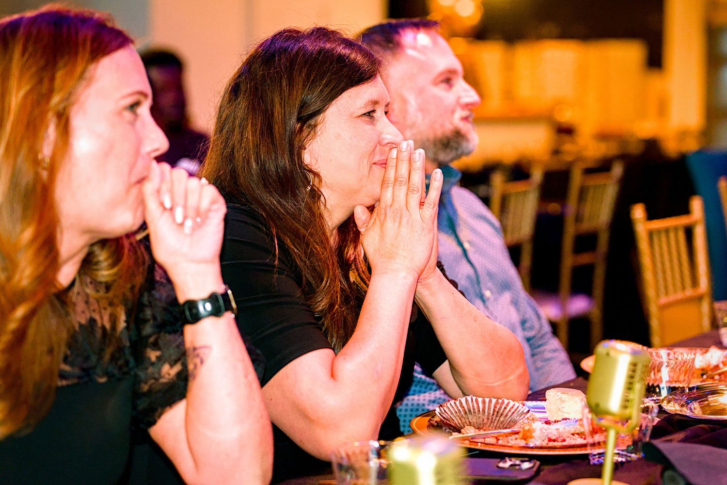 Three adults sit at a banquet table, with a woman in the center pressing her hands together in awe.