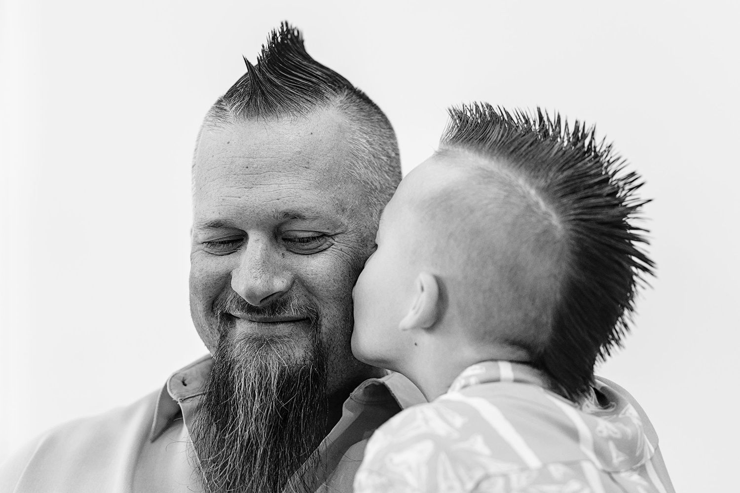 Father with mohawk and beard smiling with young child who has matching mohawk hairstyle in black and white photo.