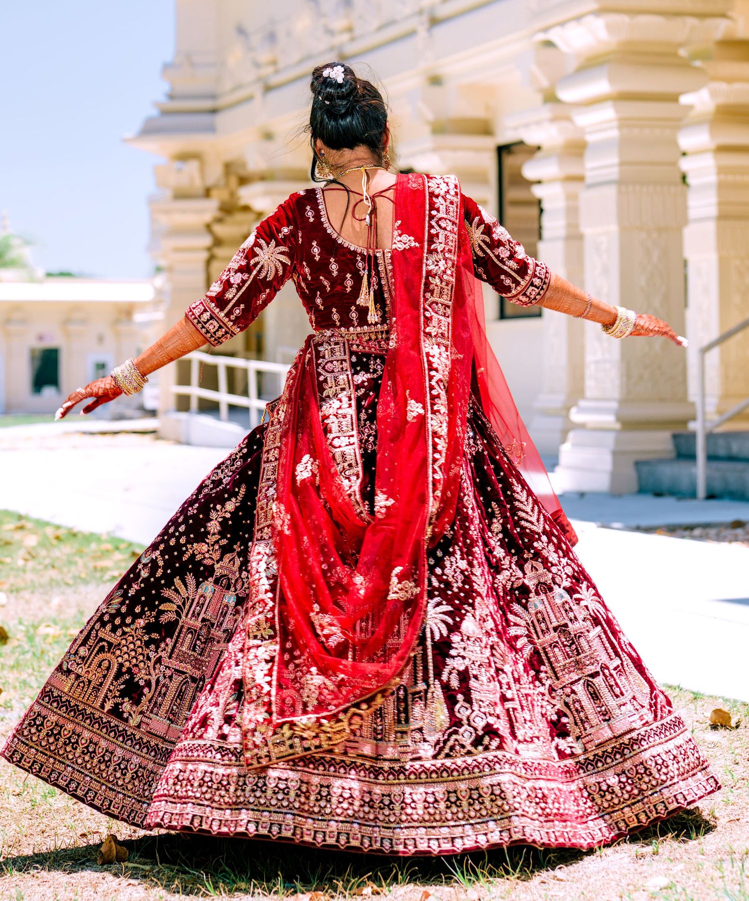 Bride twirls in stunning red and maroon embroidered bridal lehenga outside ornate temple venue.