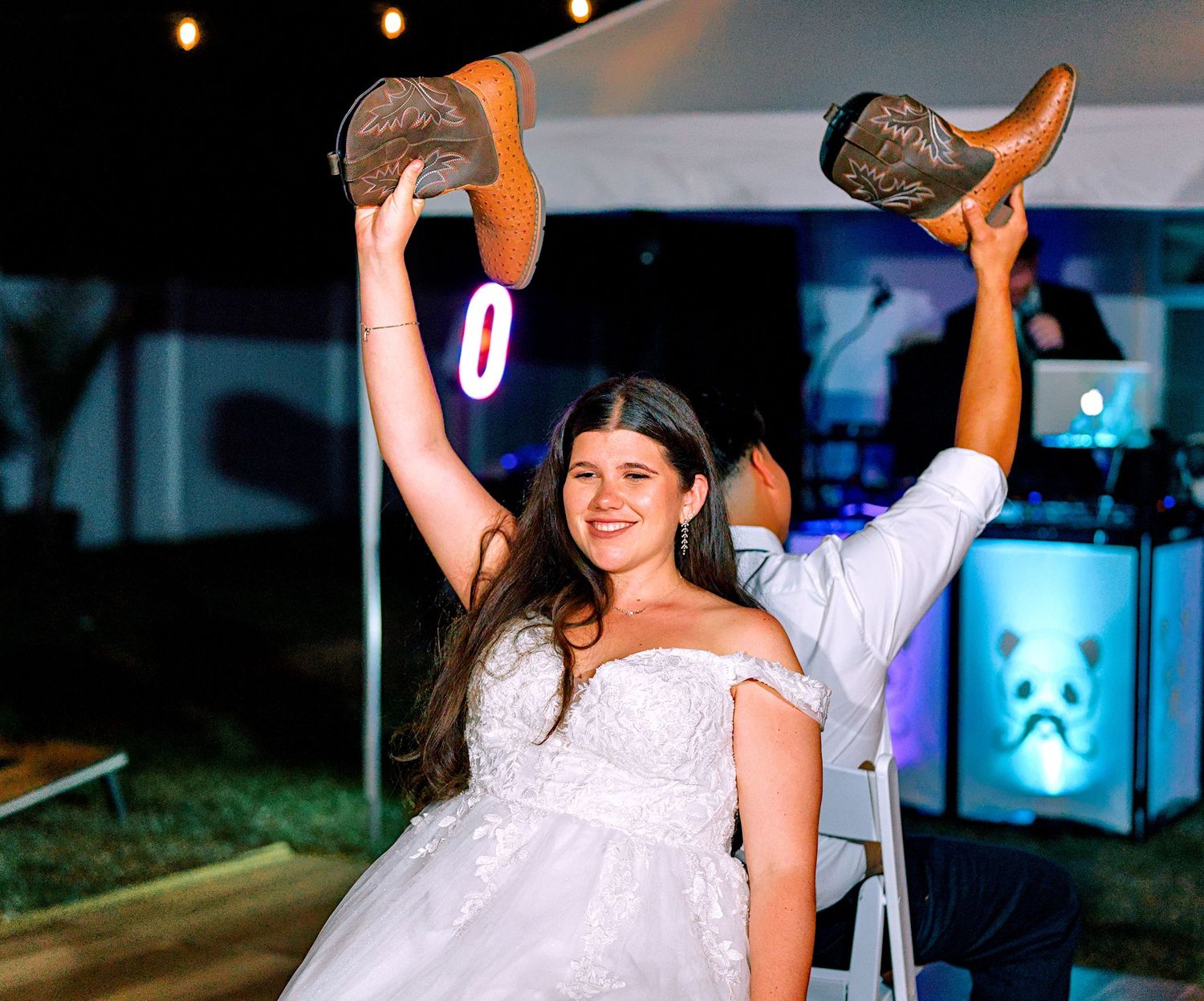 Bride in white gown holds up cowboy boots while seated in chair at outdoor wedding reception.