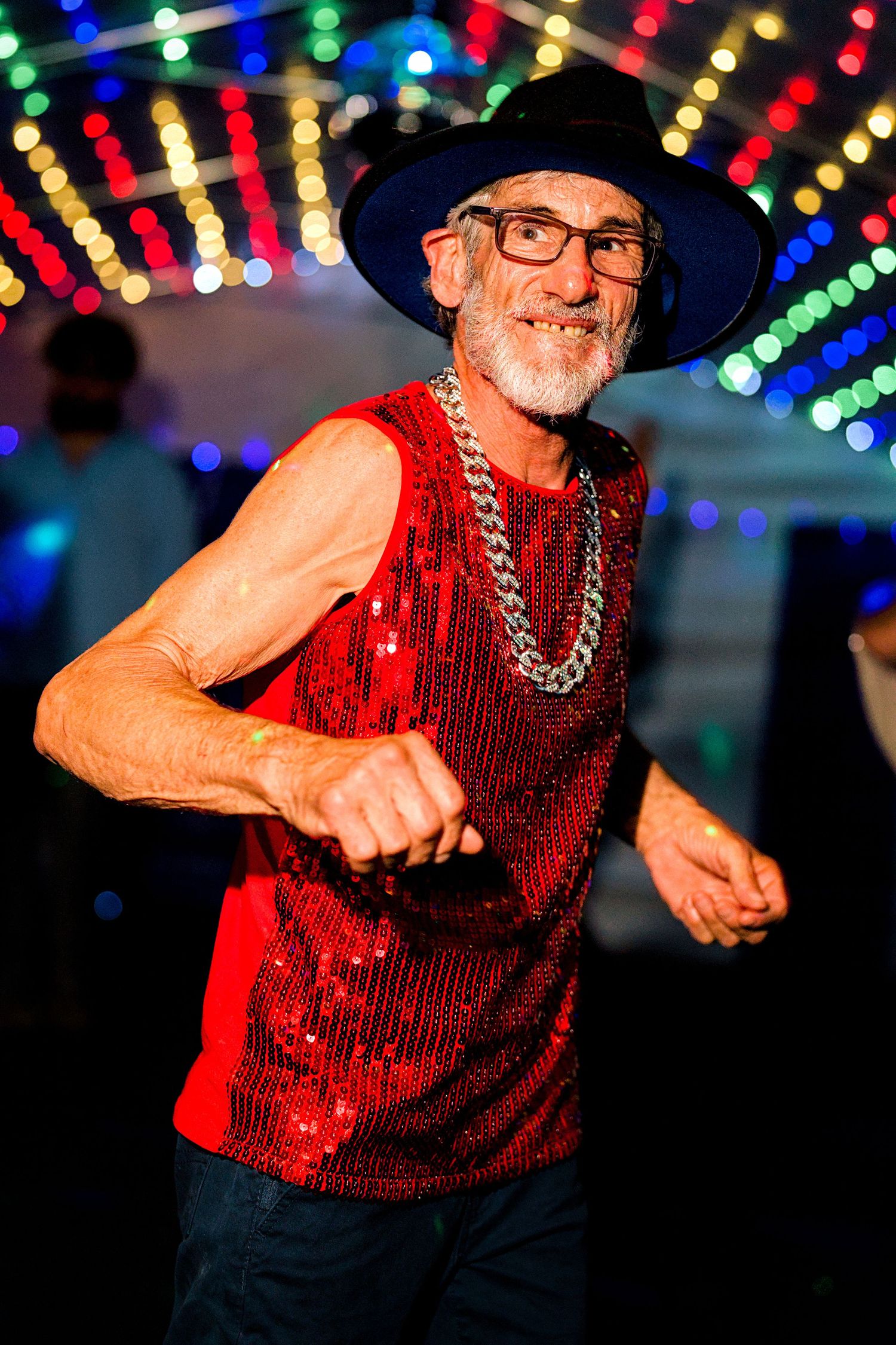 Elderly man dancing joyfully in red sequin sleeveless top, black hat, chain necklace, and glasses under colorful party lights.