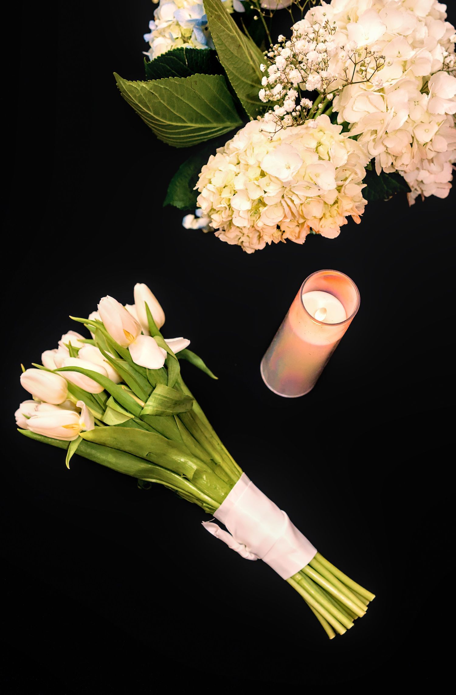A bouquet of white tulips tied with pink ribbon next to a lit candle and white hydrangeas on black background.