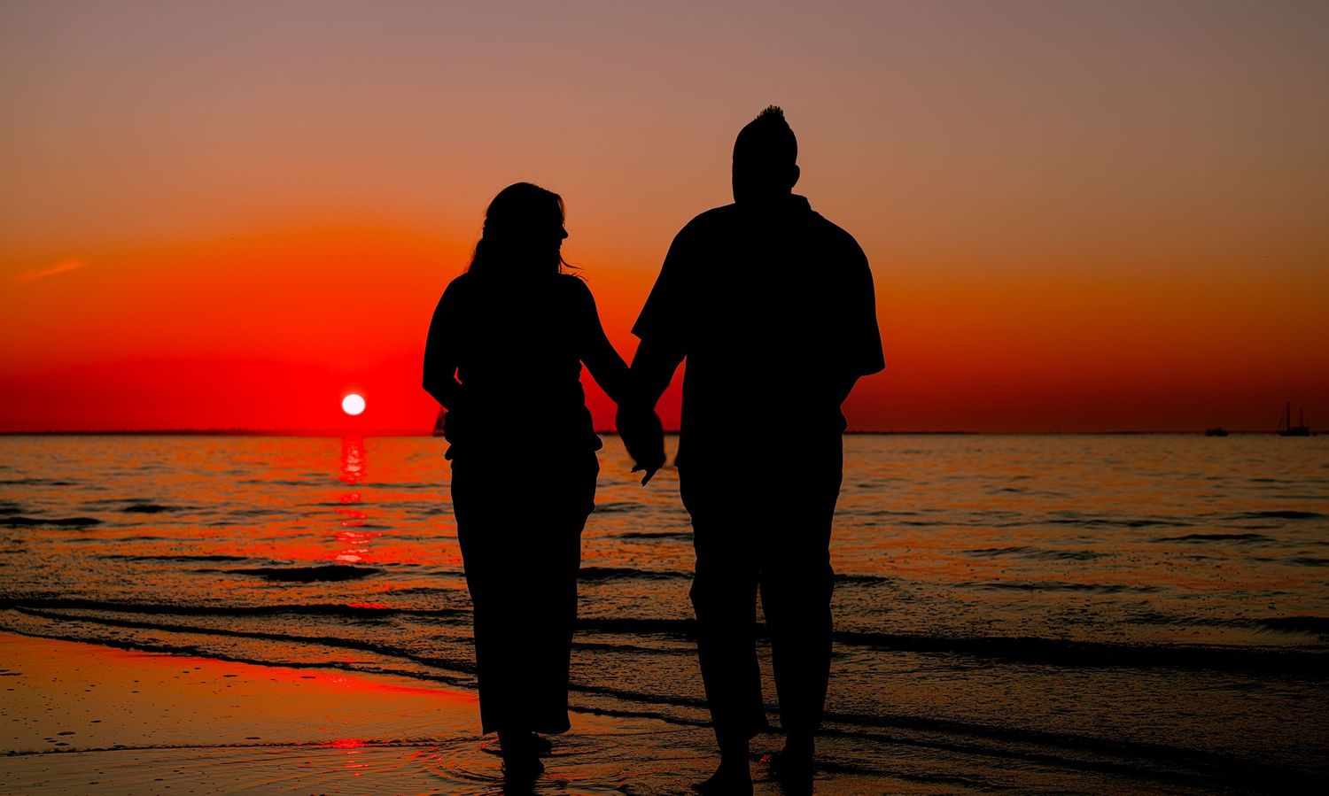 Silhouettes of people walking along a beach shoreline during a vibrant orange and red sunset over the ocean.