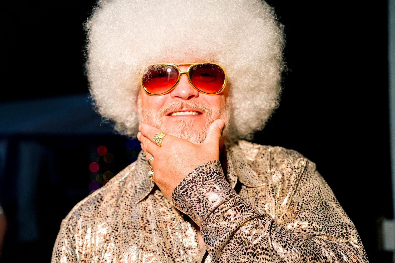 Man with white afro wig, red tinted sunglasses, and sparkly disco shirt smiling confidently.