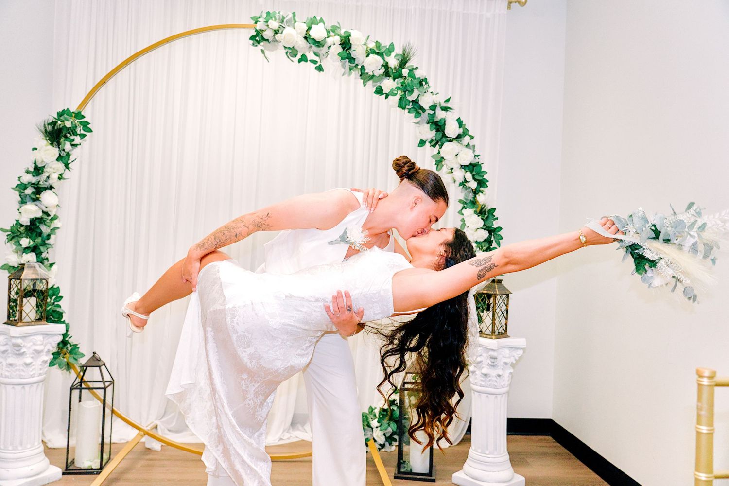 Bride and groom share a romantic dip kiss under a gold circular floral arch adorned with white roses and greenery.