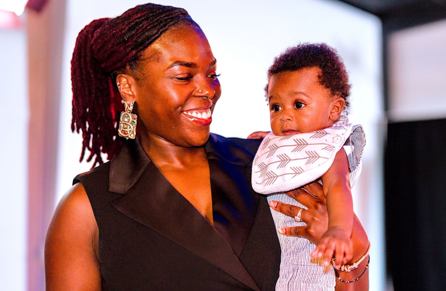 Smiling Black woman with braids holding an adorable baby at an event.