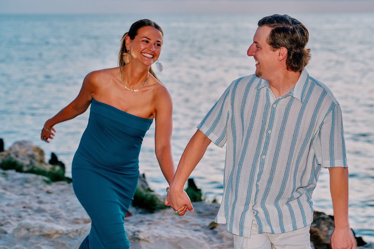 Happy couple holding hands and smiling on a rocky beach at sunset, woman in teal dress, man in striped shirt.