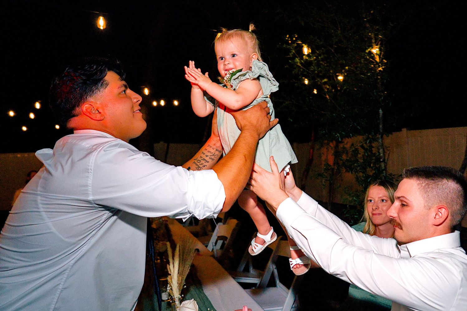 Two men joyfully lift a smiling baby girl at an outdoor evening wedding reception with string lights.