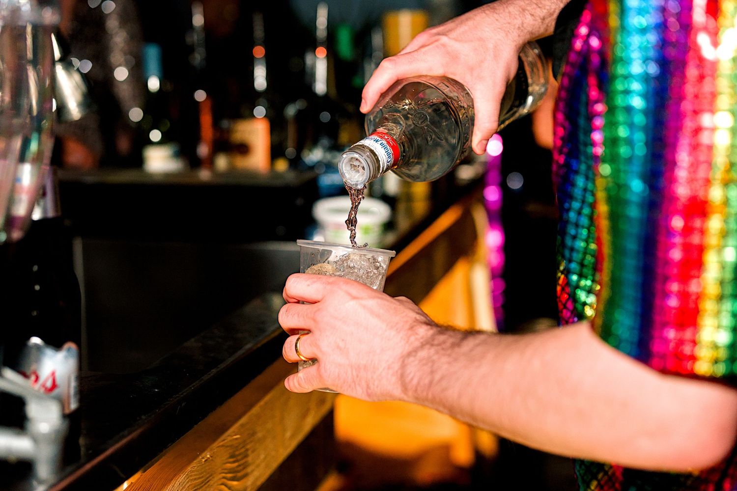 Bartender in colorful sequin shirt pouring liquor from bottle into glass at bar counter.