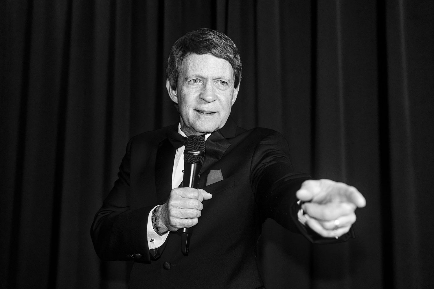 A performer in formal attire gestures dramatically during a stage performance against a dark backdrop.