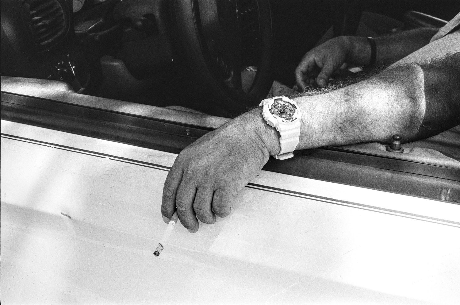 Close-up of a hand holding a cigarette out a car window with a white watch, black and white photo.