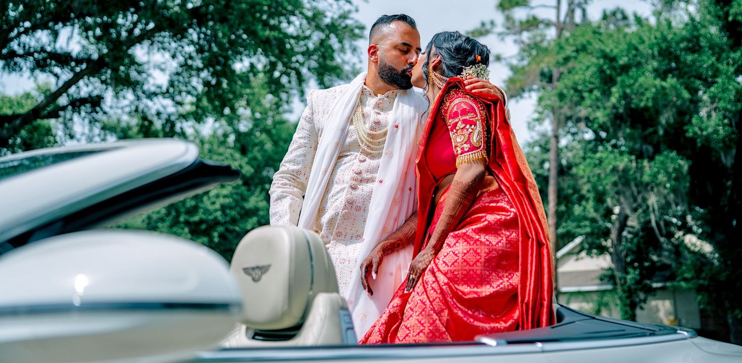 Indian couple in traditional wedding attire posing romantically by a white luxury convertible car.