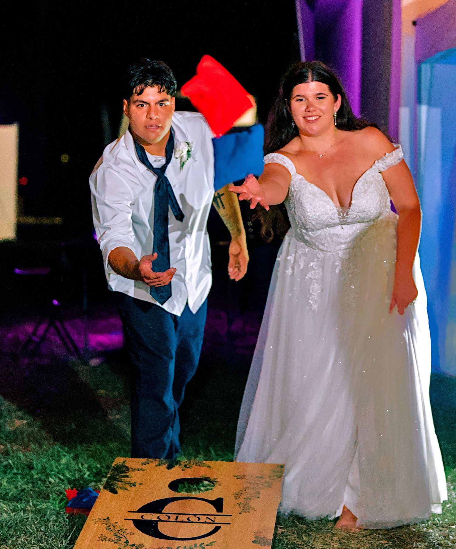 Bride and groom playing cornhole lawn game at outdoor evening wedding reception.