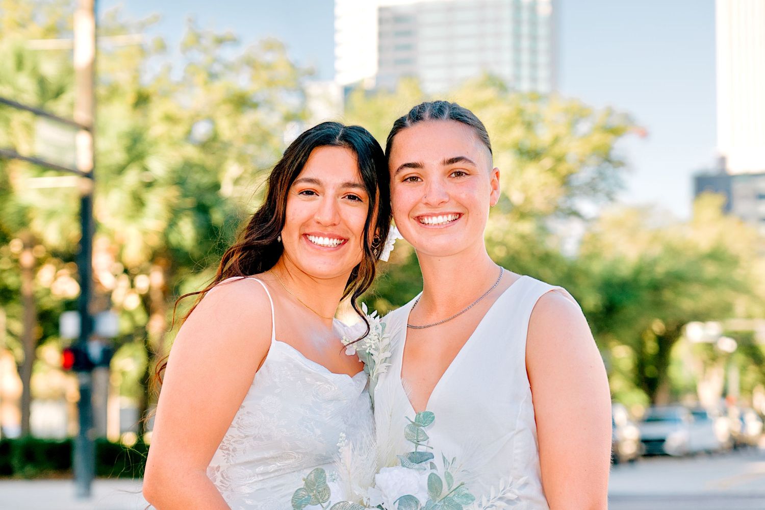 Two smiling brides in white dresses pose together on a sunny urban street with city buildings behind.