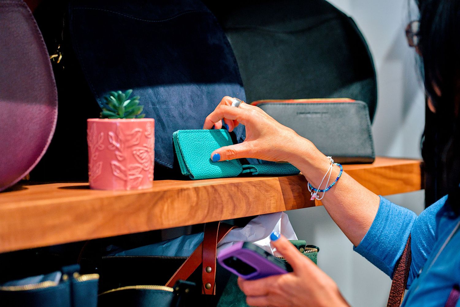 Woman picking up a teal wallet from a wooden shelf displaying colorful accessories and a small succulent plant.