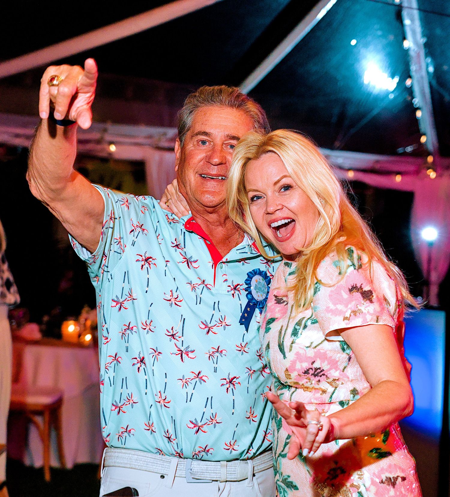 Two smiling people taking a selfie at an outdoor evening party under a tent, wearing colorful summer attire.