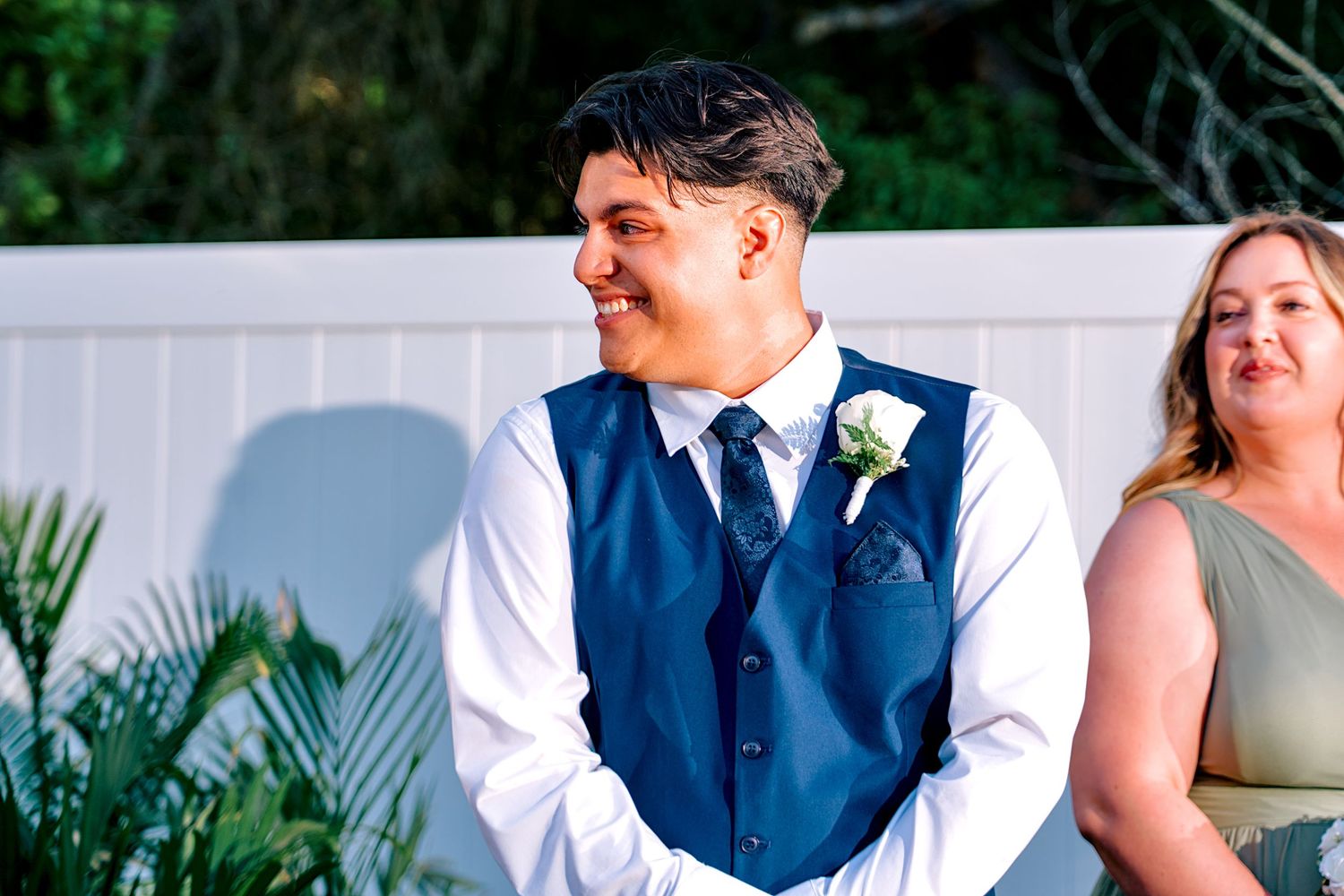 Smiling groom in blue vest and white shirt with boutonniere stands at outdoor wedding ceremony.