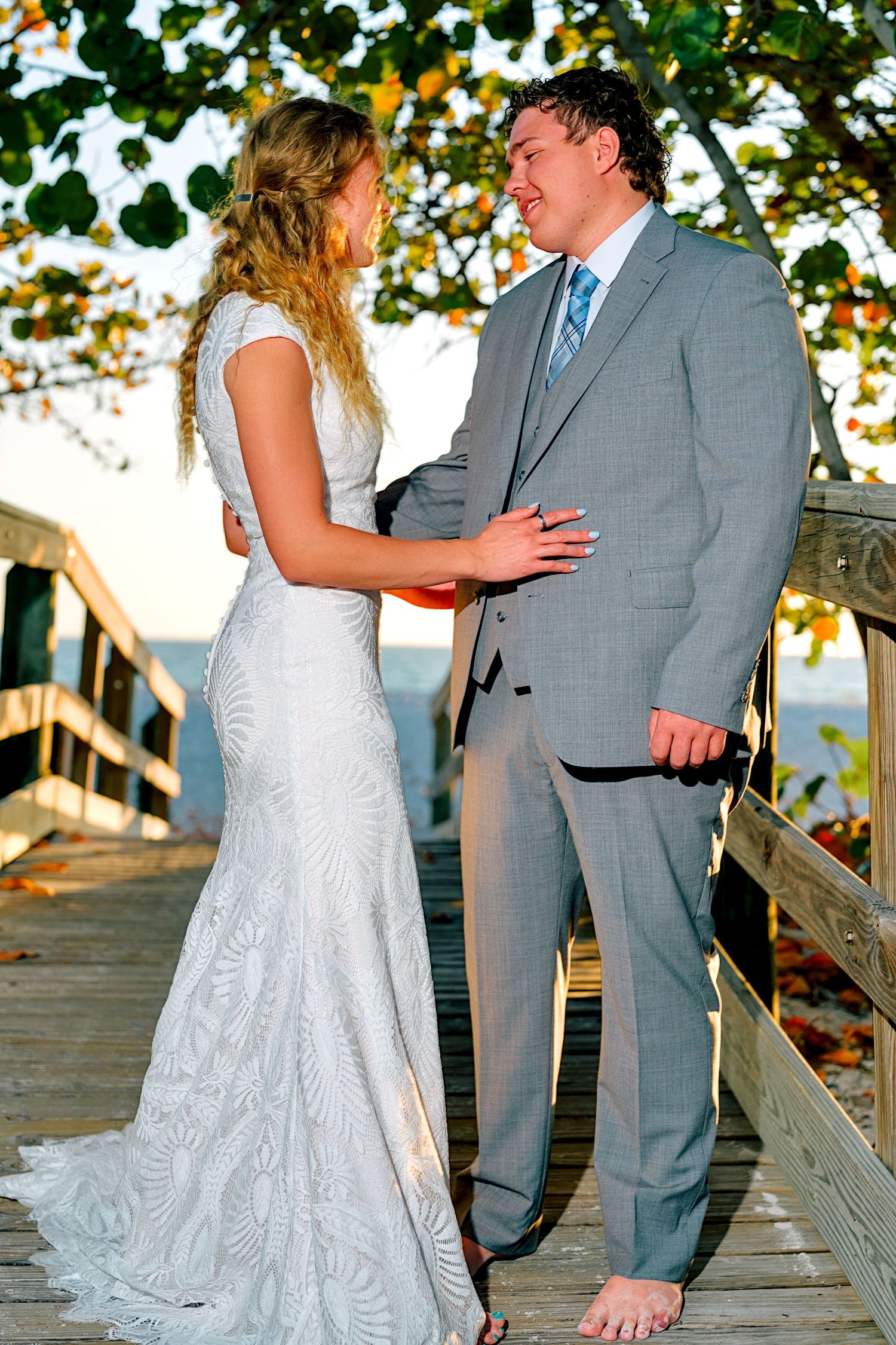 A bride in a white lace gown and groom in a gray suit share a romantic moment on a wooden boardwalk near the ocean.