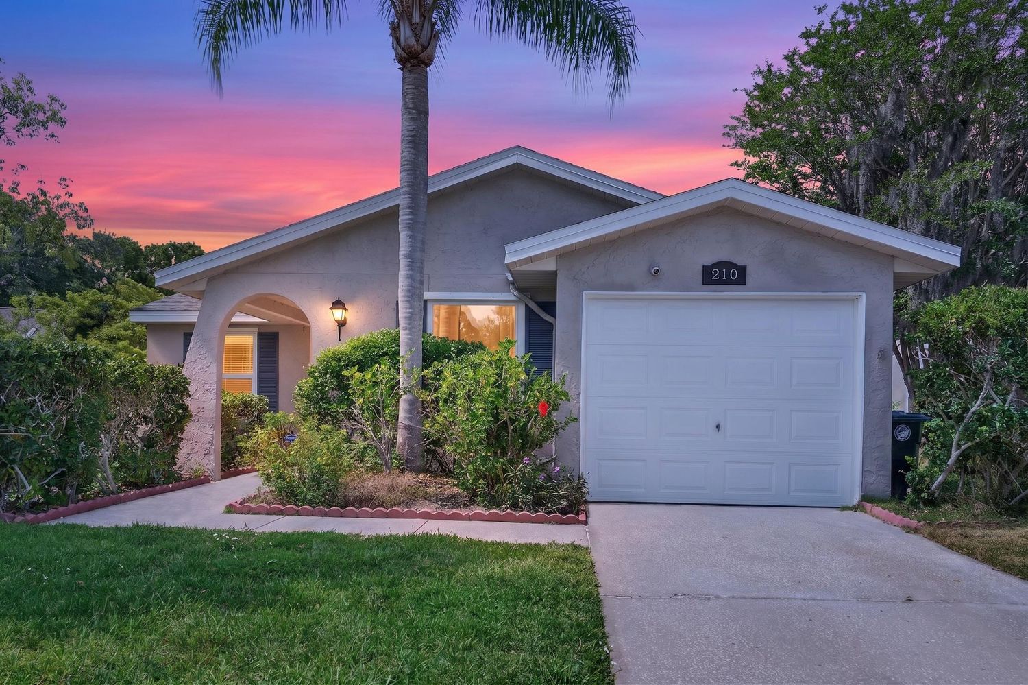 Single-story Florida home with stucco exterior, palm tree, and two-car garage under a vibrant pink and purple sunset sky.