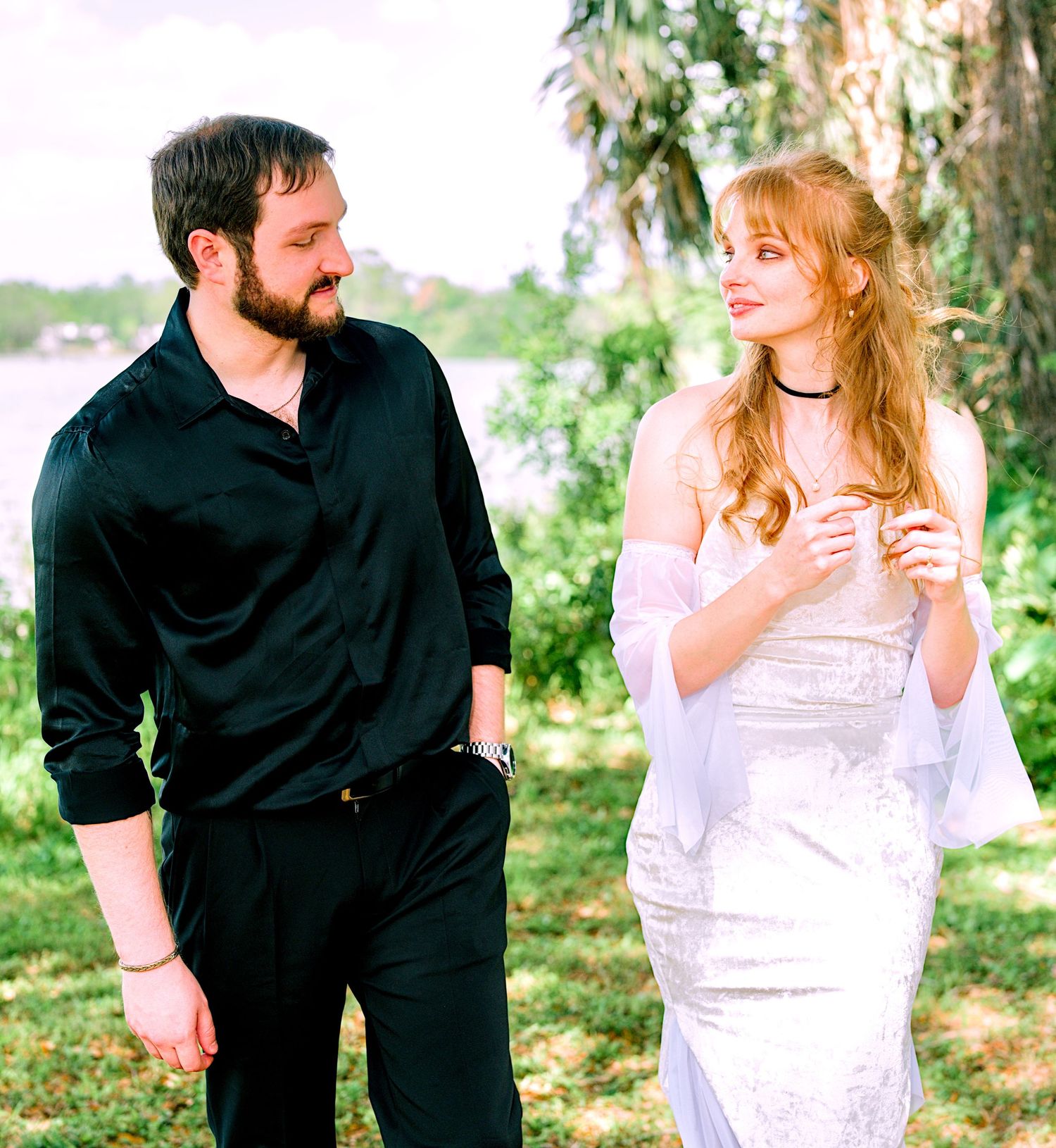 Couple walking outdoors near trees, woman in white dress and man in black outfit smiling.