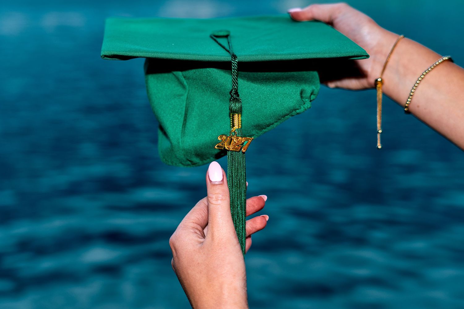 Two hands holding a green graduation cap with gold tassel against a blurred turquoise water background.