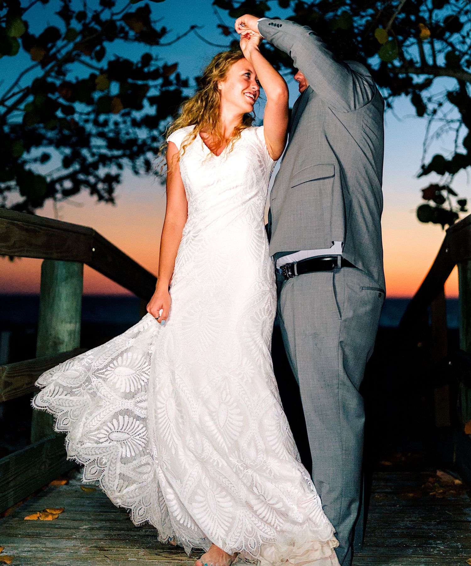 Bride and groom dancing on wooden deck at sunset during beach wedding celebration.