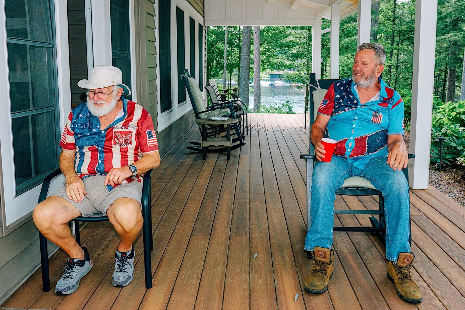 Two men relaxing in rocking chairs on a wooden porch, one wearing an American flag shirt and hat, the other in blue overalls.
