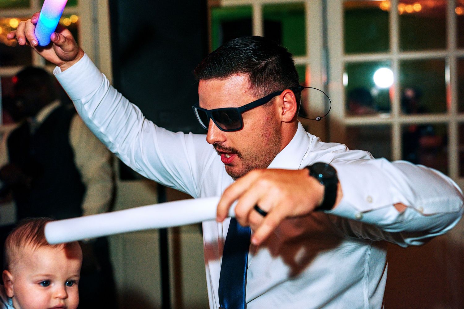 A man in a white shirt and sunglasses dances energetically with a glow stick at a wedding reception.