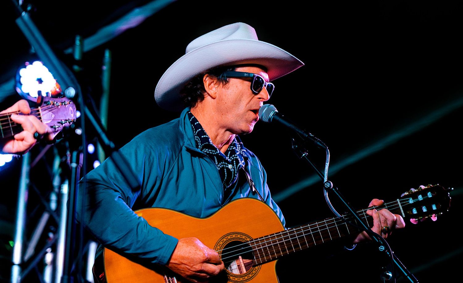 A country musician in a white cowboy hat plays acoustic guitar and sings at an outdoor concert.