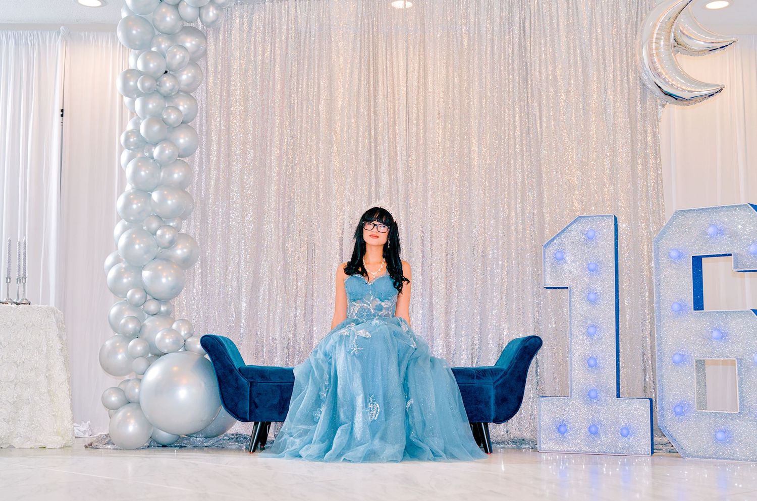 Young woman in light blue ball gown poses at Sweet 16 party with balloon decorations and large number 16.