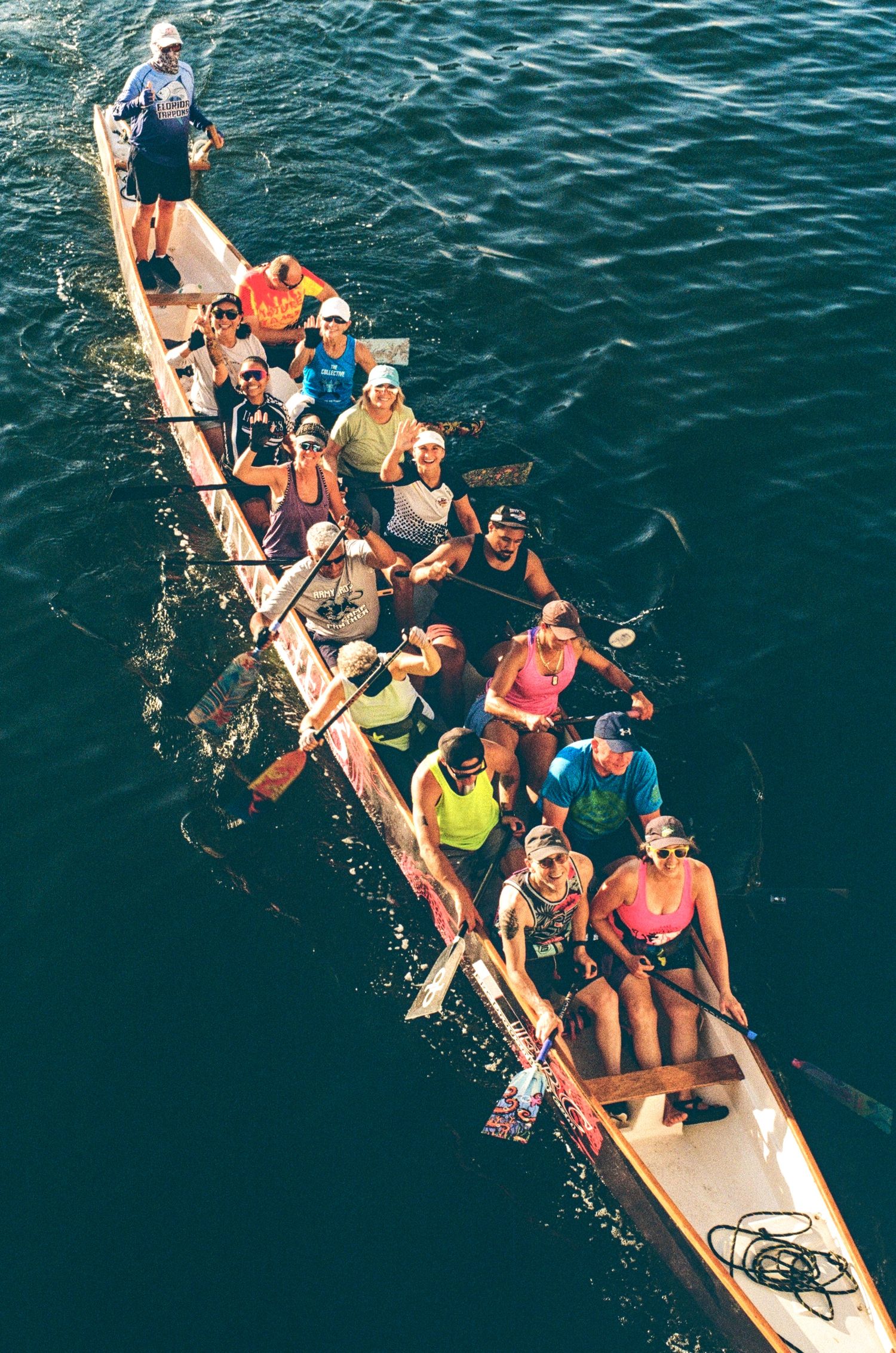 Aerial view of a large traditional outrigger canoe with a team of paddlers rowing together on calm dark blue water.