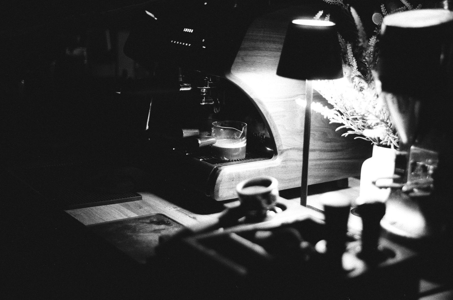 Black and white photo of a coffee machine on a cafe counter with a lamp illuminating cups nearby.